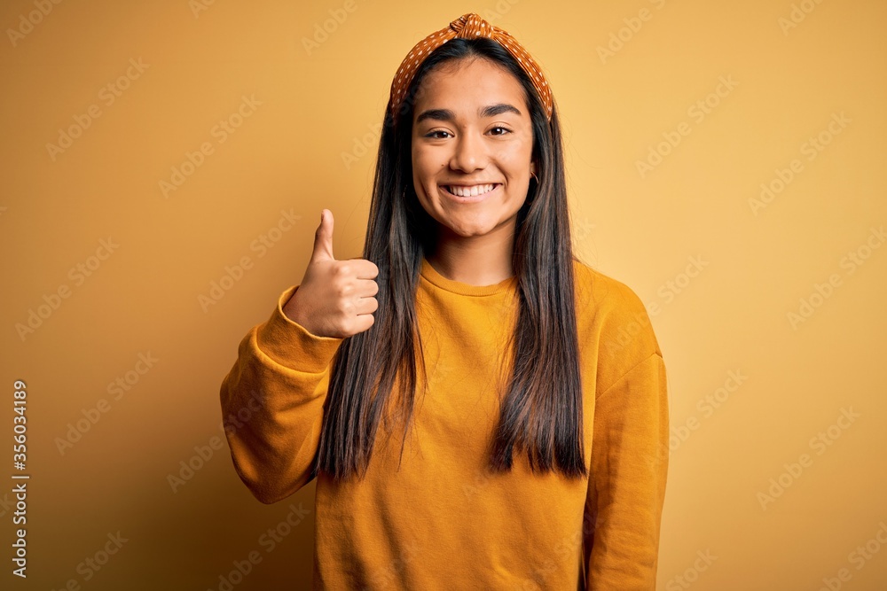 Young beautiful asian woman wearing casual sweater and diadem over yellow background doing happy thumbs up gesture with hand. Approving expression looking at the camera showing success. Young beautiful asian woman wearing casual sweater and diadem over yellow background doing happy thumbs up gesture with hand. Approving expression looking at the camera showing success.