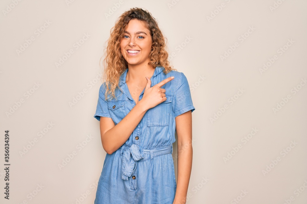 Young beautiful woman with blue eyes wearing casual denim dress over white background cheerful with a smile of face pointing with hand and finger up to the side with happy and natural expression
