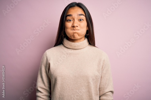 Young beautiful asian woman wearing casual turtleneck sweater over pink background puffing cheeks with funny face. Mouth inflated with air, crazy expression.