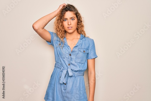 Young beautiful woman with blue eyes wearing casual denim dress over white background confuse and wonder about question. Uncertain with doubt, thinking with hand on head. Pensive concept.