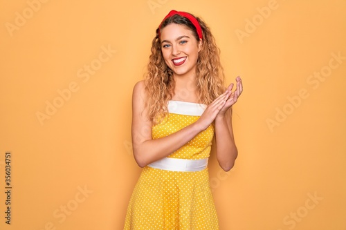 Beautiful blonde pin-up woman with blue eyes wearing diadem standing over yellow background clapping and applauding happy and joyful, smiling proud hands together