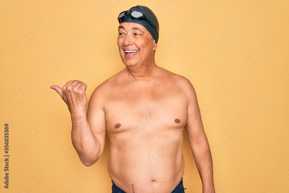 Middle age senior grey-haired swimmer man wearing swimsuit, cap and goggles smiling with happy face looking and pointing to the side with thumb up.