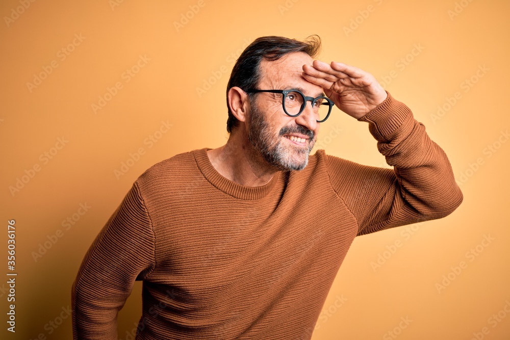 Middle age hoary man wearing brown sweater and glasses over isolated yellow background very happy and smiling looking far away with hand over head. Searching concept. Middle age hoary man wearing brown sweater and glasses over isolated yellow background very happy and smiling looking far away with hand over head. Searching concept.