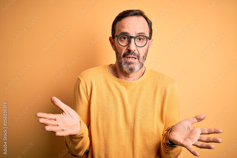 Middle age hoary man wearing casual sweater and glasses over isolated yellow background clueless and confused with open arms, no idea concept. Middle age hoary man wearing casual sweater and glasses over isolated yellow background clueless and confused with open arms, no idea concept.