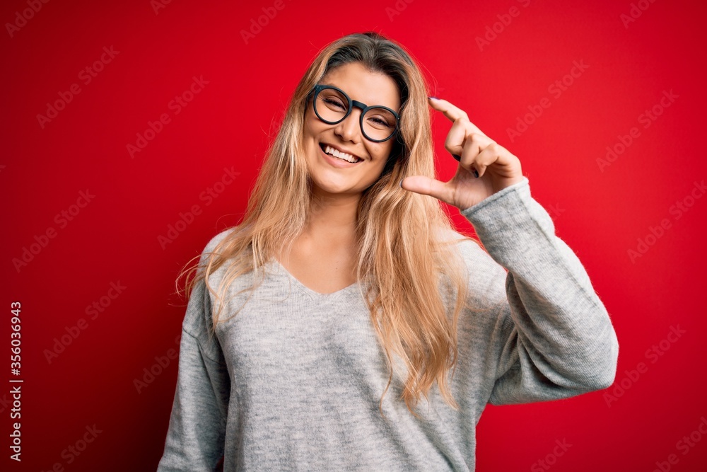Young beautiful blonde woman wearing sweater and glasses over isolated red background smiling and confident gesturing with hand doing small size sign with fingers looking and the camera. Measure