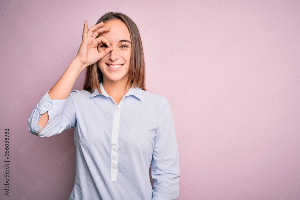 Young beautiful businesswoman wearing elegant shirt standing over isolated pink background doing ok gesture with hand smiling, eye looking through fingers with happy face. Young beautiful businesswoman wearing elegant shirt standing over isolated pink background doing ok gesture with hand smiling, eye looking through fingers with happy face.