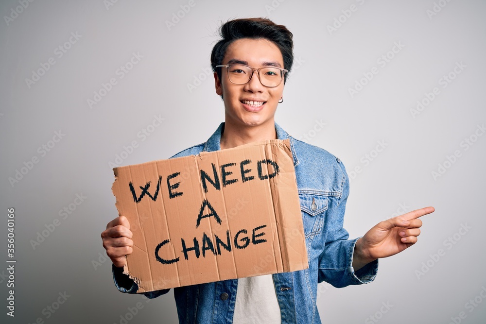 Young handsome chinese activist man protesting asking for change holding poster very happy pointing with hand and finger to the side Young handsome chinese activist man protesting asking for change holding poster very happy pointing with hand and finger to the side