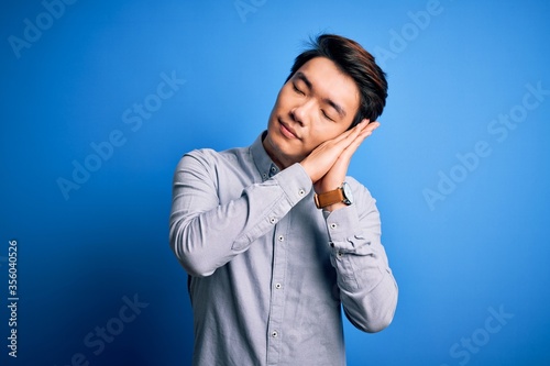 Young handsome chinese man wearing casual shirt standing over isolated blue background sleeping tired dreaming and posing with hands together while smiling with closed eyes.