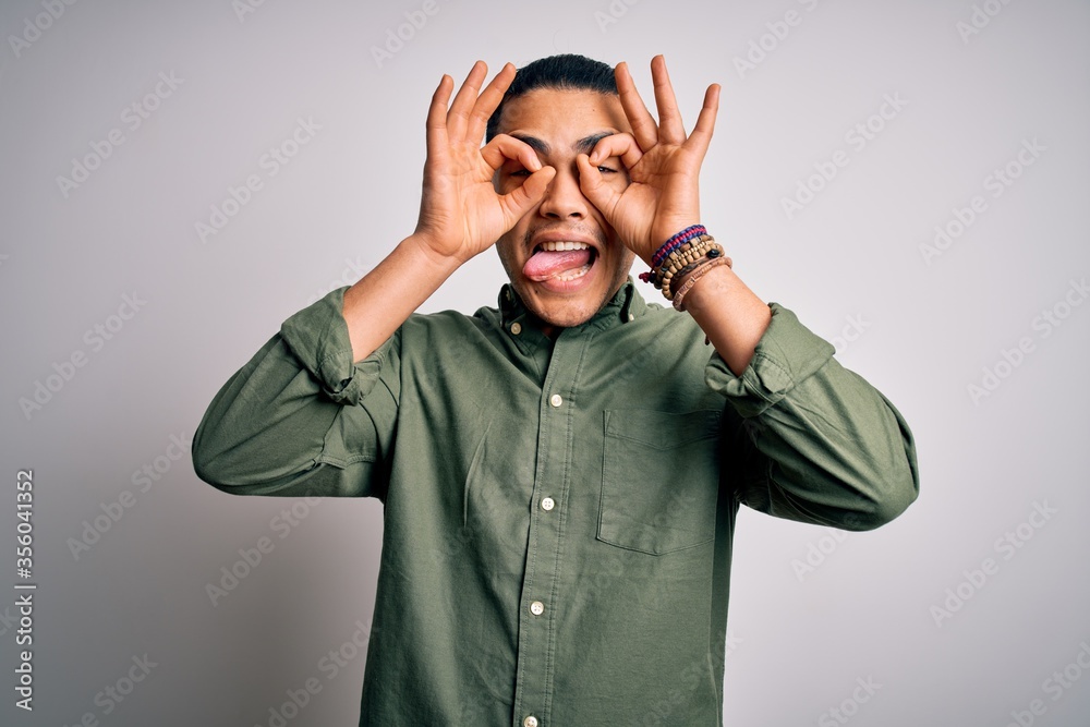 Young brazilian man wearing casual shirt standing over isolated white background doing ok gesture like binoculars sticking tongue out, eyes looking through fingers. Crazy expression. Young brazilian man wearing casual shirt standing over isolated white background doing ok gesture like binoculars sticking tongue out, eyes looking through fingers. Crazy expression.