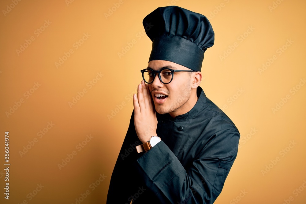 Young brazilian chef man wearing cooker uniform and hat over isolated yellow background hand on mouth telling secret rumor, whispering malicious talk conversation Young brazilian chef man wearing cooker uniform and hat over isolated yellow background hand on mouth telling secret rumor, whispering malicious talk conversation
