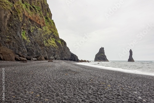 Reynisdrangar basalt rock formation in the Atlantic ocean with black sand beach and rocky mountain in overcast day typical for Iceland