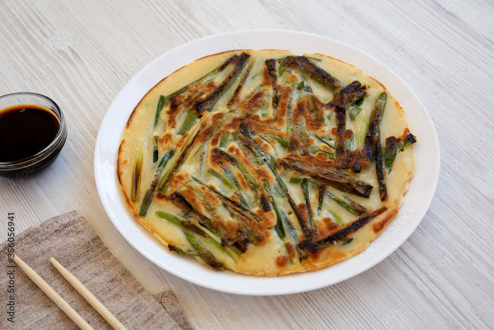 Homemade korean Pajeon scallion pancake on a white plate on a white wooden surface, side view. Asian food. Homemade korean Pajeon scallion pancake on a white plate on a white wooden surface, side view. Asian food.