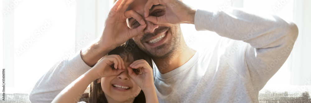 Fototapeta premium Smiling father and little daughter having fun at home, making fingers like glasses eyewear shape looking through binoculars smiling looking at camera. Horizontal photo banner for website header design