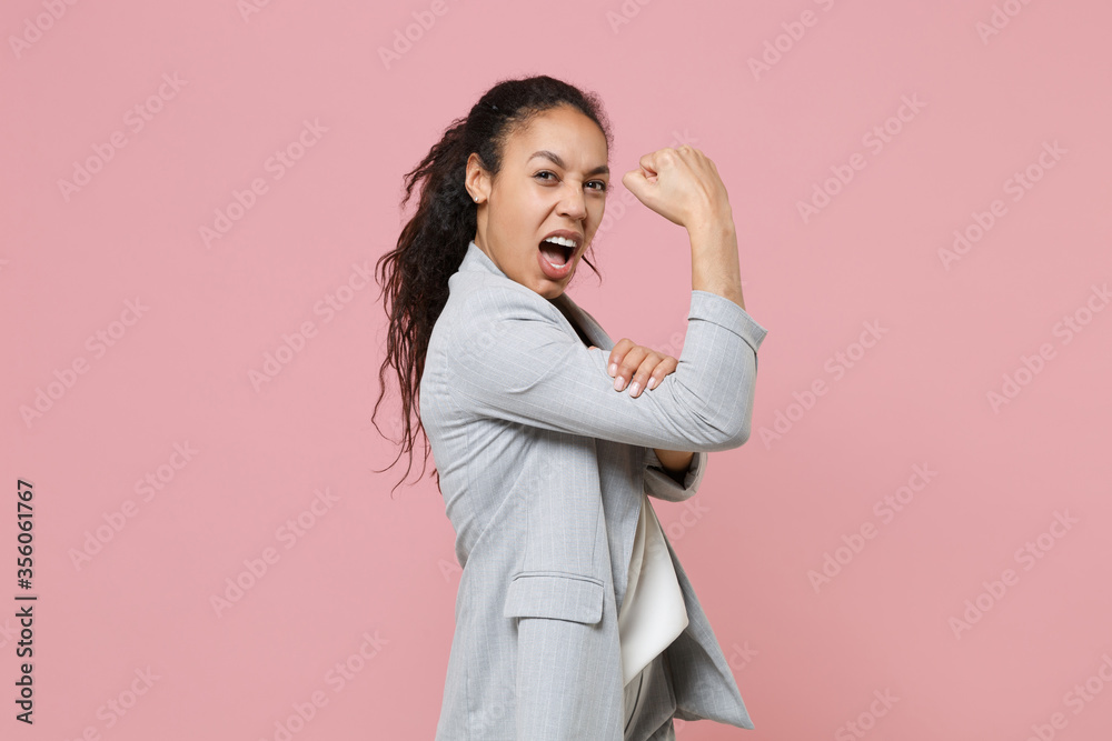 Side view of crazy young african american business woman in grey suit white shirt isolated on pink background. Achievement career wealth business concept. Mock up copy space. Showing biceps muscles. Side view of crazy young african american business woman in grey suit white shirt isolated on pink background. Achievement career wealth business concept. Mock up copy space. Showing biceps muscles.