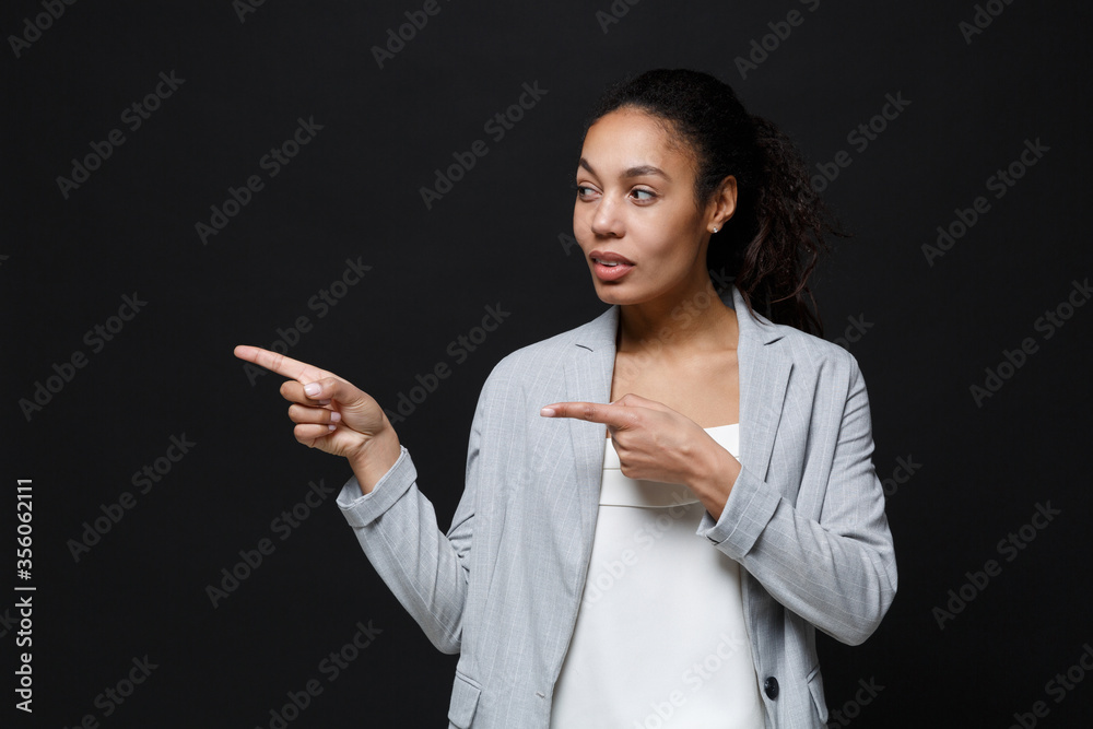 Attractive young african american business woman in grey suit, white shirt isolated on black background. Achievement career wealth business concept. Mock up copy space. Pointing index fingers aside. Attractive young african american business woman in grey suit, white shirt isolated on black background. Achievement career wealth business concept. Mock up copy space. Pointing index fingers aside.