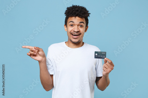 Excited young african american guy in casual white t-shirt posing isolated on blue background studio. People lifestyle concept. Mock up copy space. Hold credit bank card, pointing index finger aside.