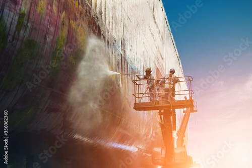 Worker Washing and Cleaning with cargo ship at side shell in floating dry dock with water jet  from sea vegetation and muse colonies during routine overhaul wearing equipment protective safety harness