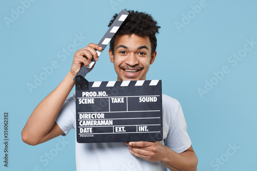 Smiling young african american guy in casual white t-shirt posing isolated on pastel blue background studio. People lifestyle concept. Mock up copy space. Hold classic black film making clapperboard.