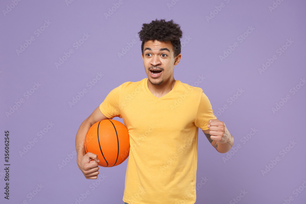 Shocked young african american guy basketball player in yellow t-shirt isolated on pastel violet background. People emotions, sport leisure lifestyle concept. Play basketball hold ball clenching fist. Shocked young african american guy basketball player in yellow t-shirt isolated on pastel violet background. People emotions, sport leisure lifestyle concept. Play basketball hold ball clenching fist.