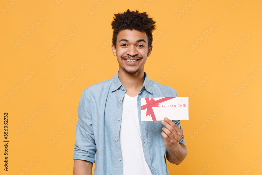 Smiling young african american guy in casual blue shirt posing isolated on yellow wall background studio portrait. People sincere emotions lifestyle concept. Mock up copy space. Hold gift certificate. Smiling young african american guy in casual blue shirt posing isolated on yellow wall background studio portrait. People sincere emotions lifestyle concept. Mock up copy space. Hold gift certificate.