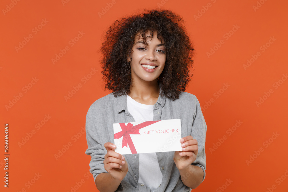 Smiling young african american woman girl in gray casual clothes isolated on orange background studio portrait. People sincere emotions lifestyle concept. Mock up copy space. Hold gift certificate. Smiling young african american woman girl in gray casual clothes isolated on orange background studio portrait. People sincere emotions lifestyle concept. Mock up copy space. Hold gift certificate.
