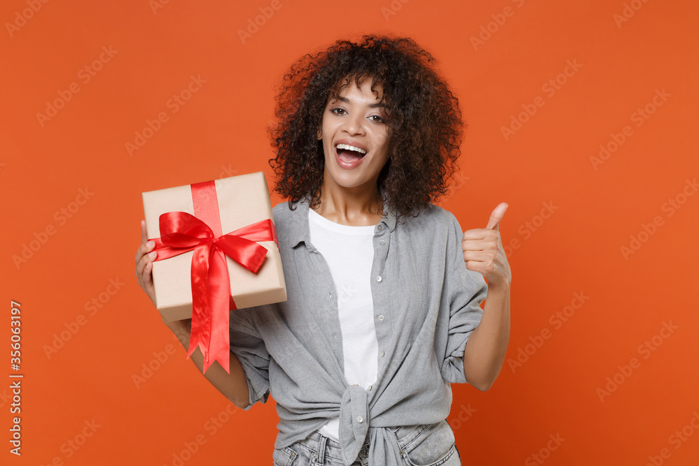 Funny young african american woman in gray casual clothes isolated on orange background. Valentine's Day Women's Day birthday, holiday concept. Hold present box with gift ribbon bow, showing thumb up. Funny young african american woman in gray casual clothes isolated on orange background. Valentine's Day Women's Day birthday, holiday concept. Hold present box with gift ribbon bow, showing thumb up.