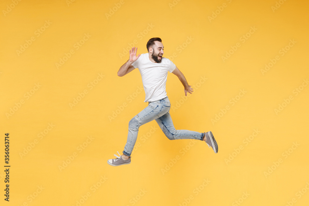 Side view of cheerful young bearded man guy in white casual t-shirt posing isolated on yellow background. People lifestyle concept. Mock up copy space. Jump like running looking aside spreading hands.