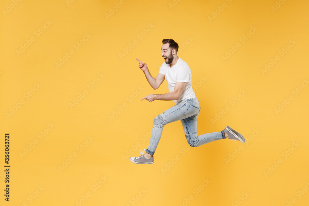 Side view of excited young bearded man guy in white casual t-shirt posing isolated on yellow wall background studio. People lifestyle concept. Mock up copy space. Jumping pointing index fingers aside.