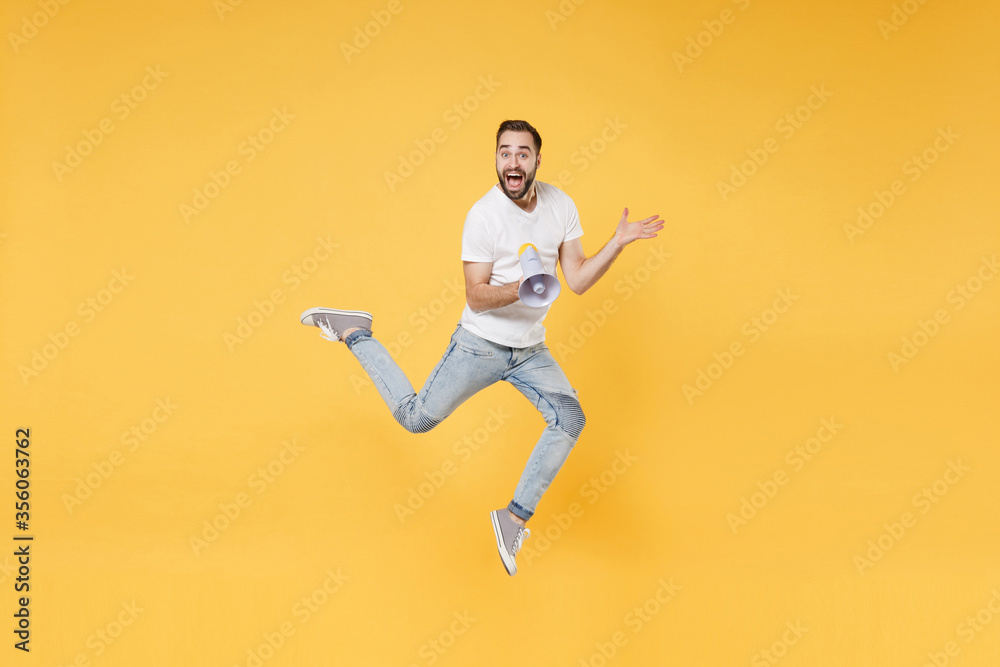 Surprised young bearded man guy in white casual t-shirt posing isolated on yellow background studio portrait. People lifestyle concept. Mock up copy space. Jumping, hold megaphone, spreading hands.
