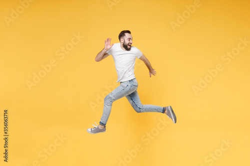 Side view of cheerful young bearded man guy in white casual t-shirt posing isolated on yellow background. People lifestyle concept. Mock up copy space. Jump like running looking aside spreading hands. © ViDi Studio