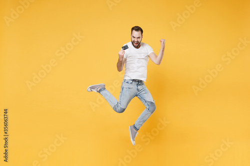 Joyful young bearded man guy in white casual t-shirt posing isolated on yellow wall background studio. People lifestyle concept. Mock up copy space. Jumping hold credit bank card doing winner gesture. © ViDi Studio