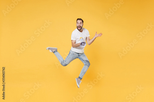 Surprised young bearded man guy in white casual t-shirt posing isolated on yellow background studio portrait. People lifestyle concept. Mock up copy space. Jumping, hold megaphone, spreading hands.