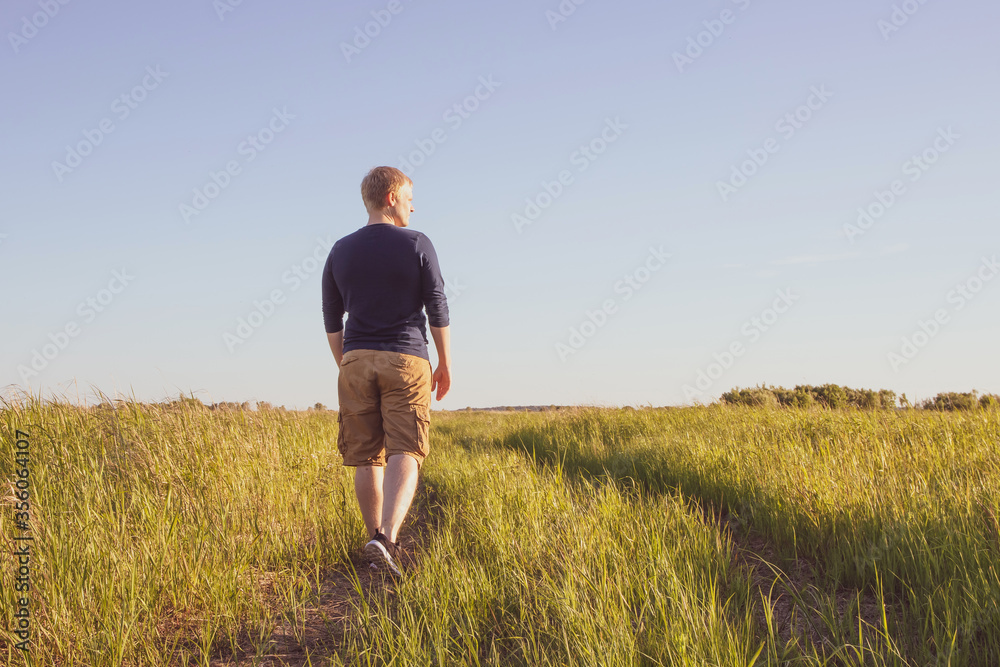 The concept of walking alone. A young Caucasian blond man walks along a path in a meadow on a sunny day. A cozy look in calm warm colors. The concept of walking alone. A young Caucasian blond man walks along a path in a meadow on a sunny day. A cozy look in calm warm colors.