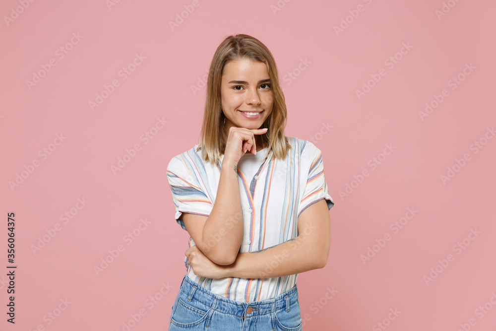 Smiling young blonde woman in casual striped shirt posing isolated on pastel pink background studio portrait. People sincere emotions lifestyle concept. Mock up copy space. Put hand prop up on chin.