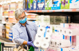 Senior woman wearing mask and rubber gloves pushes the trolley in the supermarket, reading information on the packing of the toilet paper, active elderly pensioners.