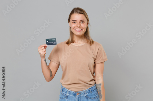 Smiling young blonde woman girl in casual beige t-shirt posing isolated on gray wall background studio portrait. People sincere emotions lifestyle concept. Mock up copy space. Hold credit bank card.