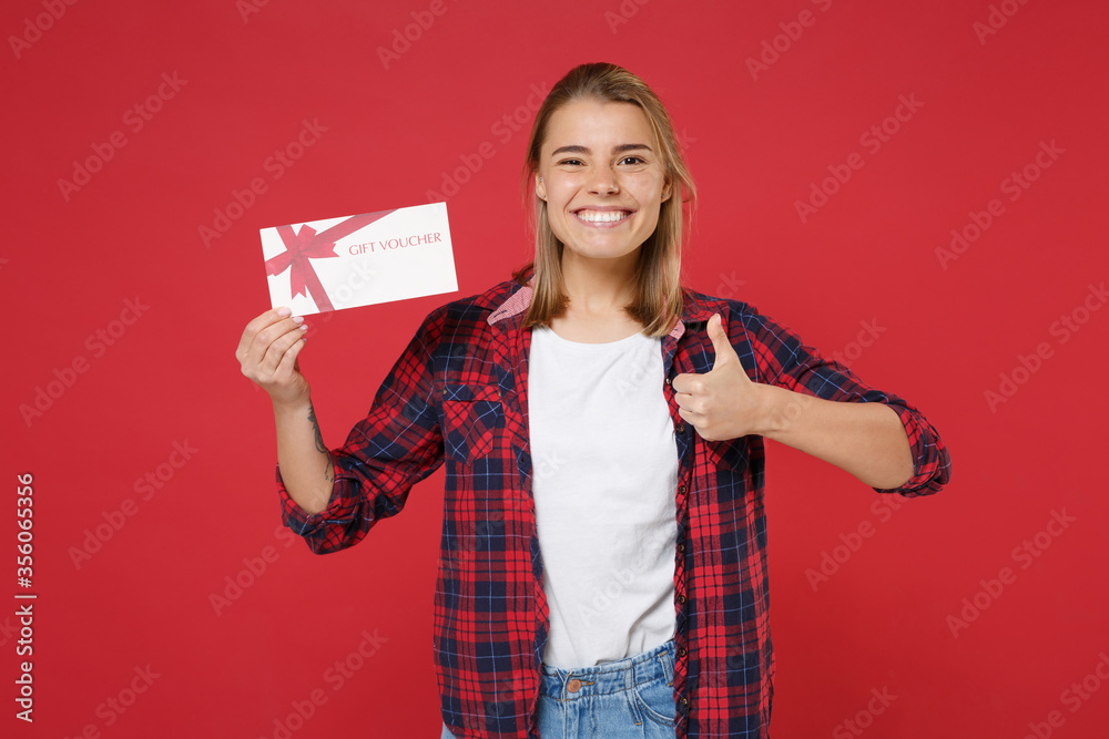 Smiling young blonde woman girl in casual checkered shirt posing isolated on red wall background studio portrait. People lifestyle concept. Mock up copy space. Hold gift certificate, showing thumb up. Smiling young blonde woman girl in casual checkered shirt posing isolated on red wall background studio portrait. People lifestyle concept. Mock up copy space. Hold gift certificate, showing thumb up.