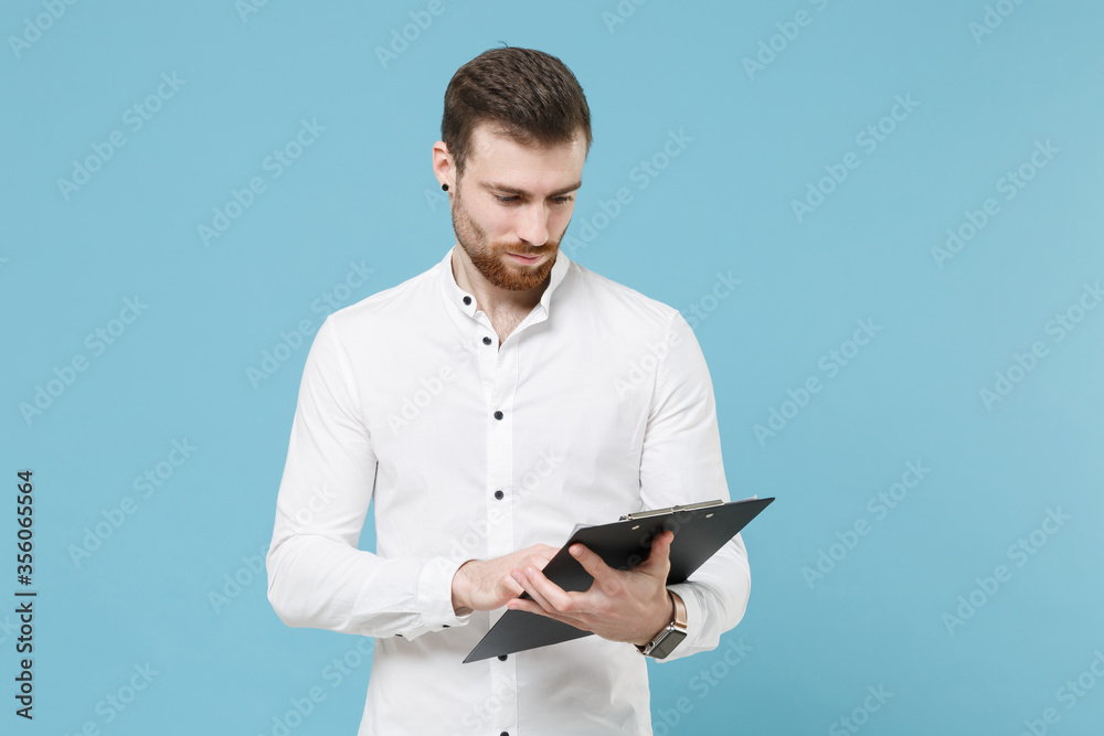 Handsome young bearded man guy 20s in white classic shirt isolated on pastel blue wall background studio portrait. People lifestyle concept. Mock up copy space. Hold clipboard with papers document.