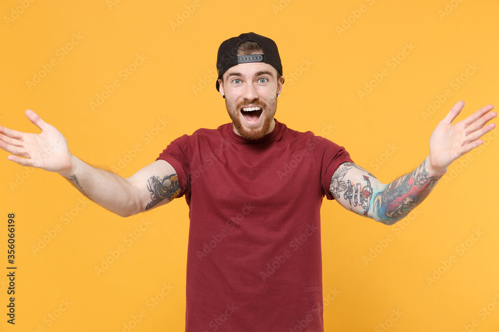 Excited young bearded tattooed man guy in casual t-shirt, black cap posing isolated on yellow wall background. People lifestyle concept. Mock up copy space. Standing with outstretched hands for hugs.