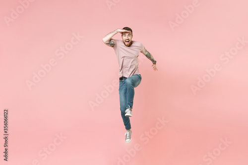Excited young bearded tattooed man guy in pastel casual t-shirt isolated on pink background. People lifestyle concept. Mock up copy space. Jumping, holding hand at forehead looking far away distance.