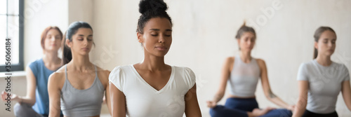 Multi ethnic pretty women seated cross-legged do meditation practice during group work out yoga session at sport club lead by african female trainer. Horizontal photo banner for website header design