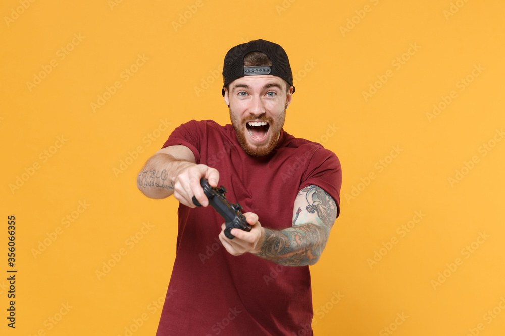 © ViDi Studio - Excited young bearded tattooed man guy in casual t-shirt black cap posing isolated on yellow wall background studio portrait. People lifestyle concept. Mock up copy space. Play game with joystick. © ViDi Studio - Excited young bearded tattooed man guy in casual t-shirt black cap posing isolated on yellow wall background studio portrait. People lifestyle concept. Mock up copy space. Play game with joystick.