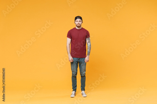 Handsome young tattooed man guy in casual t-shirt black cap posing isolated on yellow wall background studio portrait. People sincere emotions lifestyle concept. Mock up copy space. Looking camera.