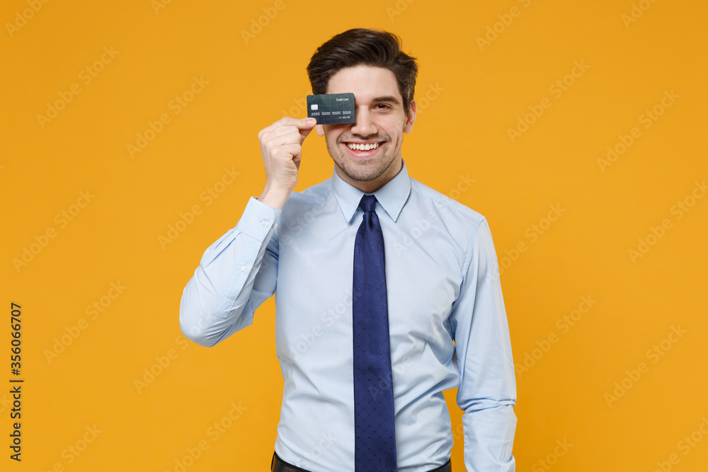 Smiling young business man in classic blue shirt tie posing isolated on yellow background studio. Achievement career wealth business concept. Mock up copy space. Covering eye with credit bank card. Smiling young business man in classic blue shirt tie posing isolated on yellow background studio. Achievement career wealth business concept. Mock up copy space. Covering eye with credit bank card.