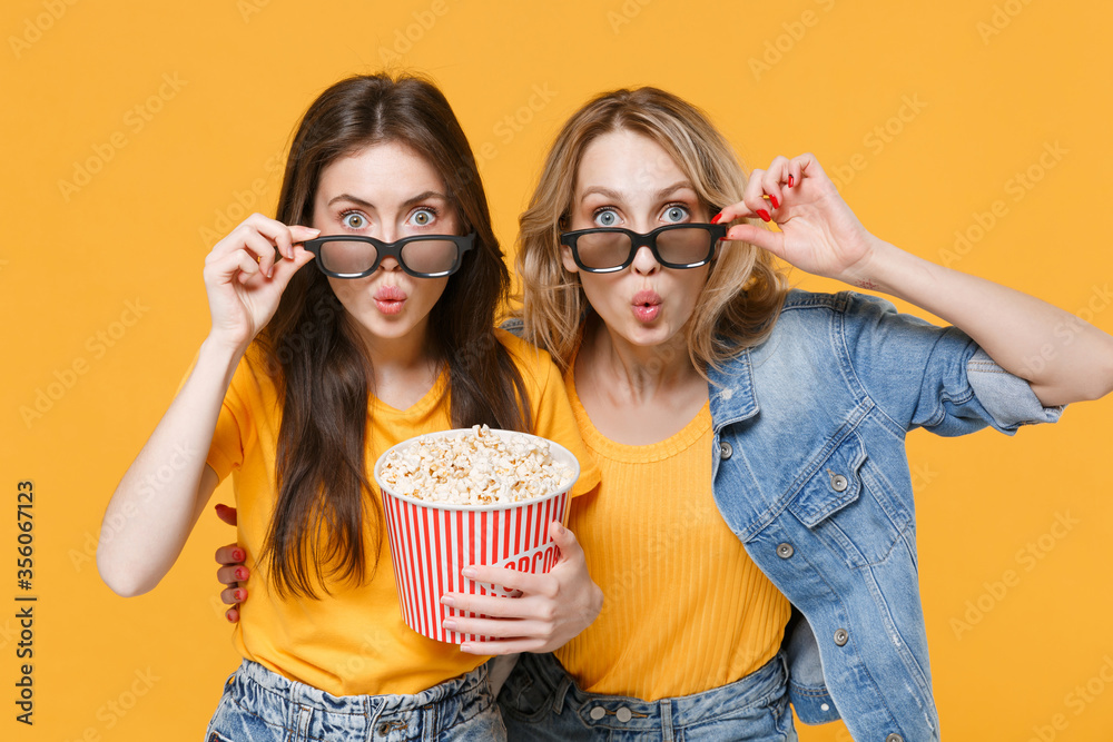 Shocked amazed young women girls friends in denim clothes, 3d glasses isolated on yellow background. People sincere emotions in cinema lifestyle concept. Watching movie film hold bucket of popcorn.