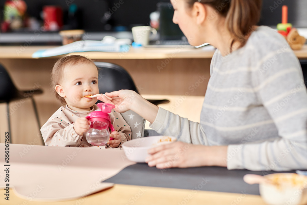 family, eating and people concept - happy mother with puree and spoon feeding little baby girl sitting in highchair at home family, eating and people concept - happy mother with puree and spoon feeding little baby girl sitting in highchair at home