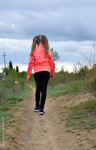 girl goes on a dirt road. green grass on the sides, cloudy gray-blue sky above the girl