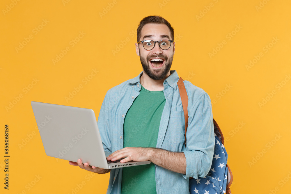 Excited young man student in casual clothes glasses backpack isolated on yellow wall background. Education in high school university college concept. Mock up copy space. Working on laptop pc computer. Excited young man student in casual clothes glasses backpack isolated on yellow wall background. Education in high school university college concept. Mock up copy space. Working on laptop pc computer.