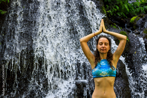 Hands raising up in namaste mudra. Young woman meditating  practicing yoga and pranayama with namaste mudra near waterfall. Yoga outdoor concept. Pucak Manik waterfall Wanagiri  Bali.