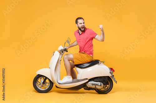 Side view of angry young bearded man guy in summer clothes driving moped isolated on yellow background. Driving motorbike transportation concept. Mock up copy space. Clenching fist screaming swearing. © ViDi Studio